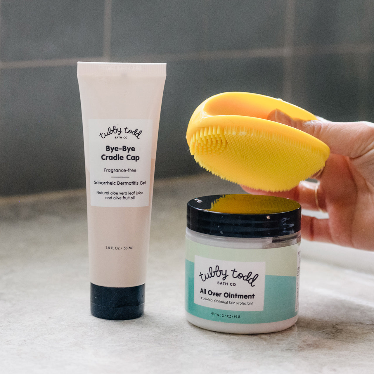 Hand holding yellow silicon brush next to Bye-Bye Cradle Cap and All Over Ointment on sink